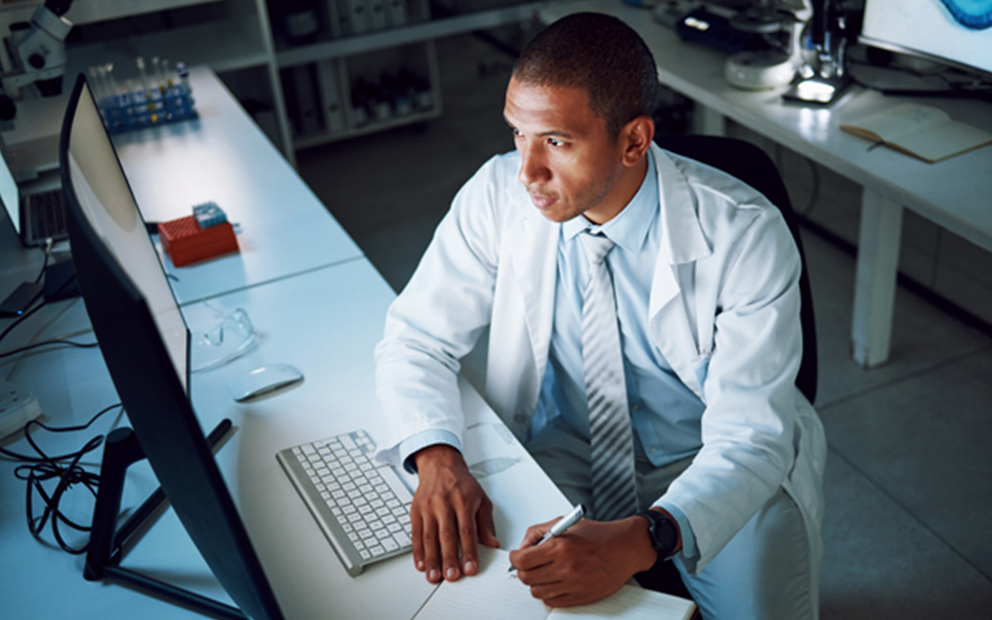 male in lab coat sitting at a computer writing in a notebook